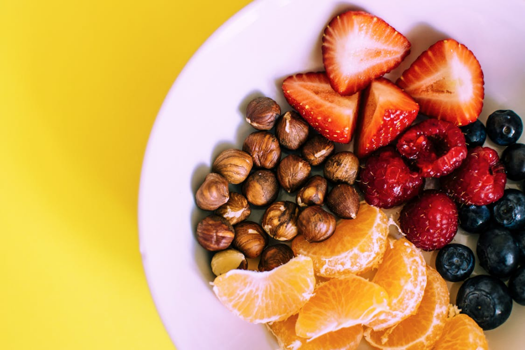 Healthiest snacks plate with colorful fresh fruits including strawberries, raspberries, blueberries, orange segments, and a cluster of hazelnuts on a yellow and white background.