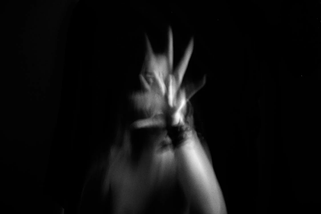 Blurred black and white photograph creating an abstract, ghostly effect. The image shows what appears to be outstretched hands or possibly a face in motion, creating a sense of distortion and movement against a dark background.