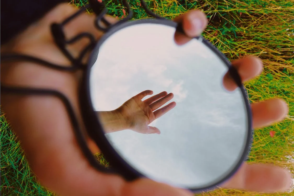 A hand holding a circular mirror outdoors against green grass. The mirror reflects the sky and clouds, creating a visual portal that contrasts with the surrounding landscape. The reflection creates an alternative perspective of reality.
