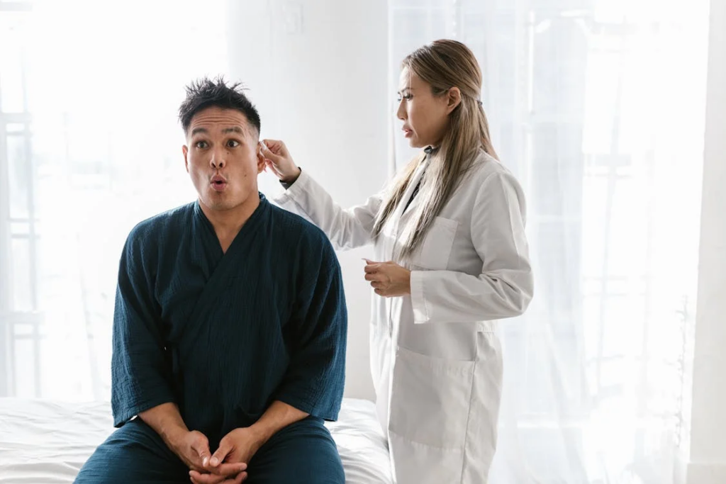A medical examination scene in a bright clinical setting. A female doctor in a white coat is examining a male patient wearing a blue medical gown who sits on an examination table with a surprised expression as the doctor checks his ear.