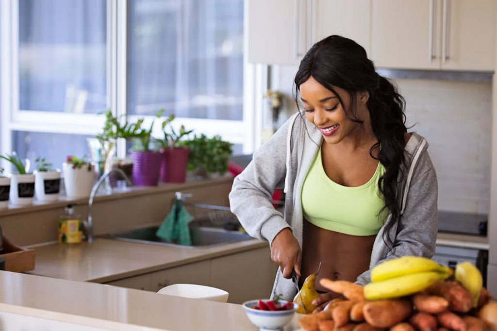 A smiling woman in a light green sports bra and gray hoodie preparing food in a kitchen. She's cutting fruit next to a counter with fresh produce including bananas and root vegetables. Plants in colorful pots are visible on the windowsill behind her.