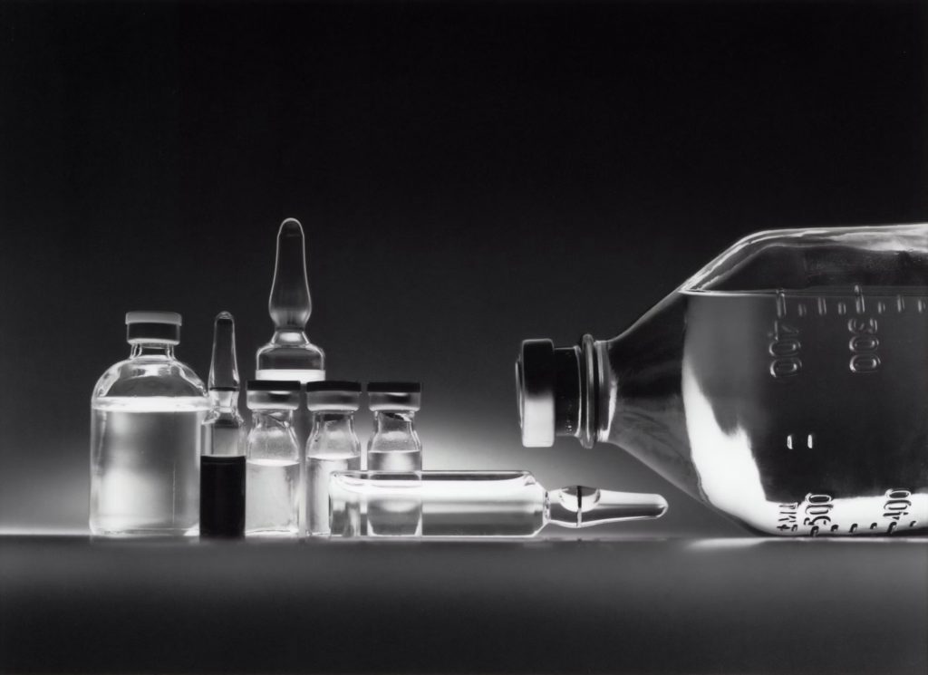 A black and white photograph showing various medical vials, ampoules, and a syringe arranged next to a larger IV solution bottle. The image displays different sizes of pharmaceutical containers against a dark background, creating a stark contrast that highlights the transparent glass and liquid contents.