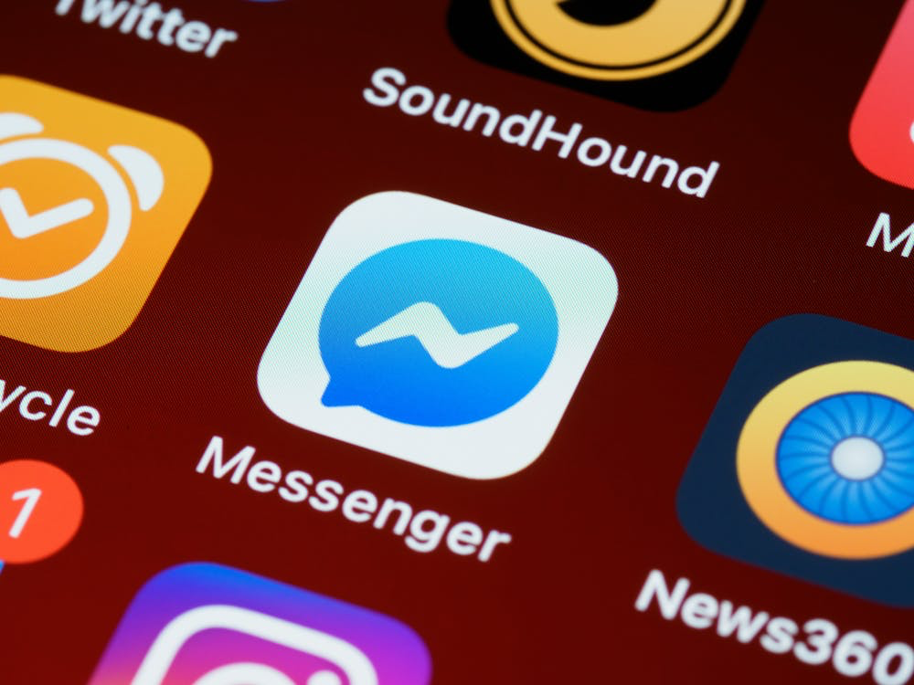 Close-up of Facebook Messenger app icon displayed on a smartphone screen alongside other social media and communication apps. The blue Messenger logo with white lightning bolt symbol is prominently visible against a dark red background, representing the platform where screenshotting Facebook chats has become a privacy concern.
