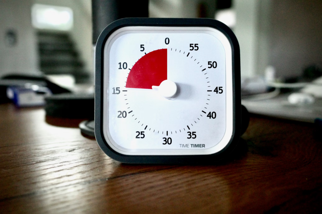 A small desk timer with a minimalist design featuring a white face and black frame. The timer displays a red section indicating elapsed time (approximately 15 minutes) with clear minute markings around the dial. The timer sits on a wooden desk surface with blurred office items in the background.