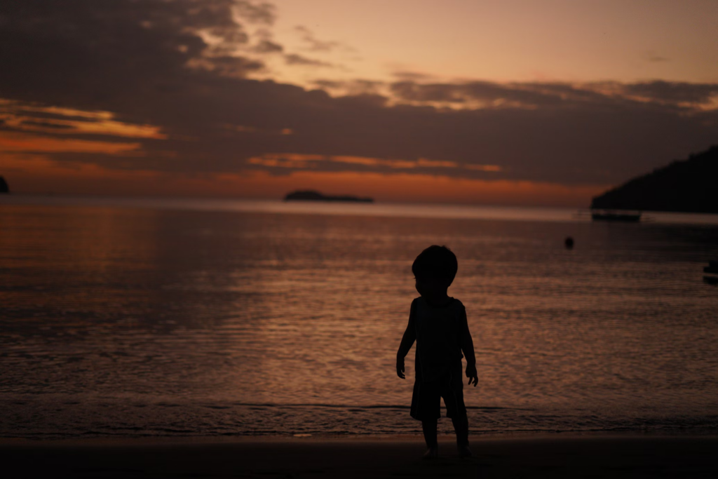 Silhouette of a small child standing at the water's edge during sunset, with orange and purple sky reflecting on calm water, creating a mysterious, contemplative mood.