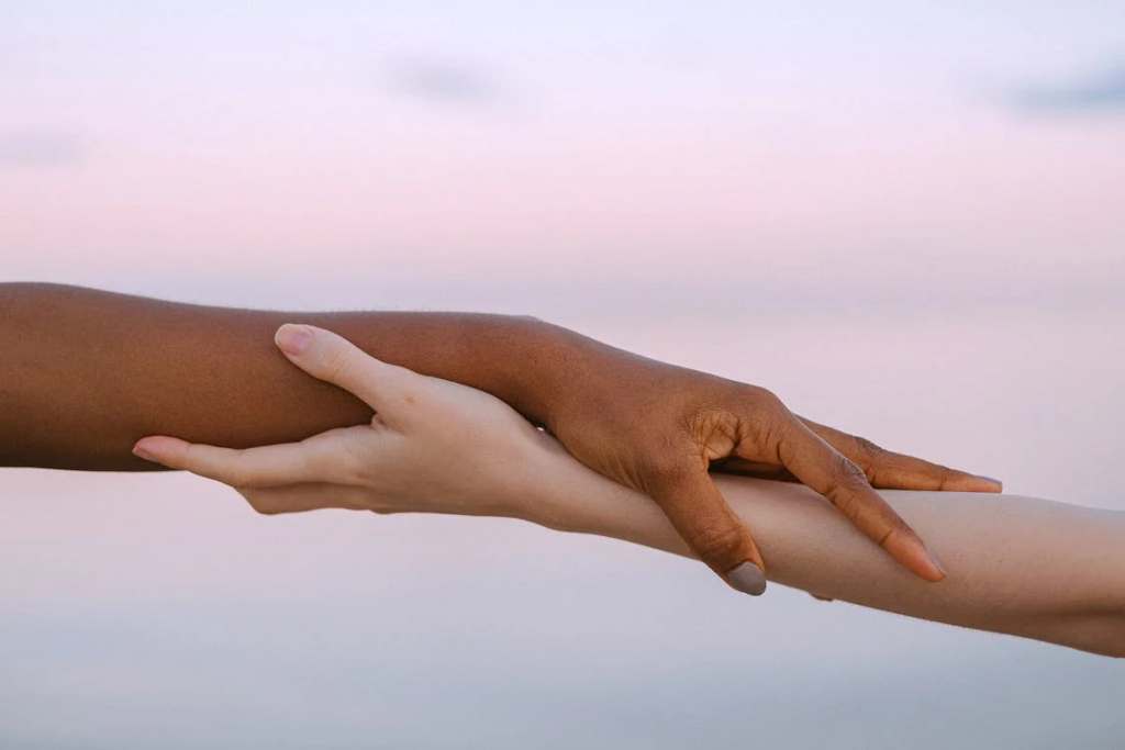 Two hands reaching toward each other against a soft pink sky - one darker-skinned and one lighter-skinned - with fingers gently touching in a connection reminiscent of Michelangelo's Creation of Adam.