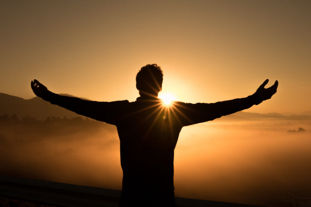 Silhouette of a person with arms outstretched against a golden sunset, standing on a mountaintop with fog below, sunlight streaming through their form in a moment of triumph or liberation.