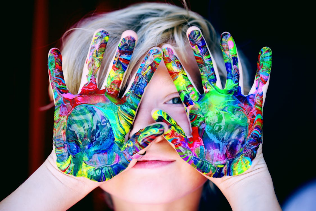 Child with vibrant rainbow-colored paint covering both hands held up in front of their face, showing bright blues, reds, yellows and greens in a playful expression of creativity and imagination.