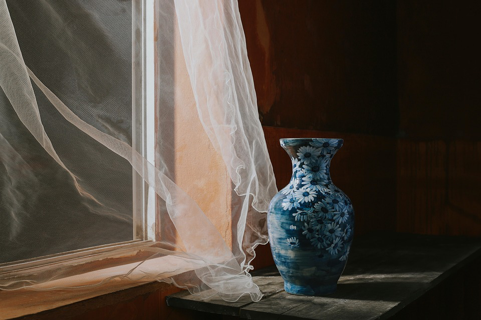 Delicate sheer curtain billowing gently beside a window, with a blue and white porcelain flower vase sitting in warm sunlight, creating a serene, peaceful domestic scene.