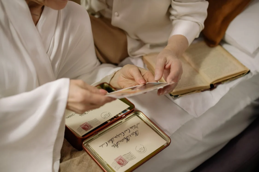 Close-up of hands in white clothing examining old photographs and letters on what appears to be a bed, with vintage correspondence box and photo album visible, suggesting someone revisiting cherished memories.
