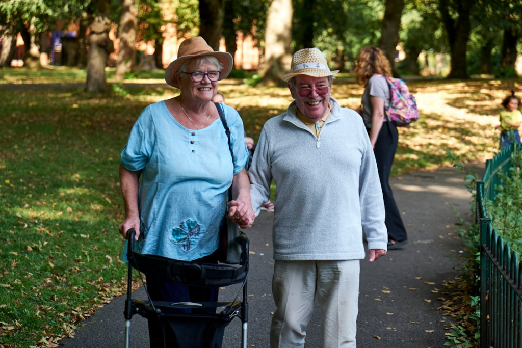 Two senior women smiling and walking together on a park path. The woman on the left wears a blue top and brown hat, using a walker for mobility support. The woman on the right wears a light-colored outfit with a straw hat. They're holding hands while walking through a tree-lined park with dappled sunlight. 