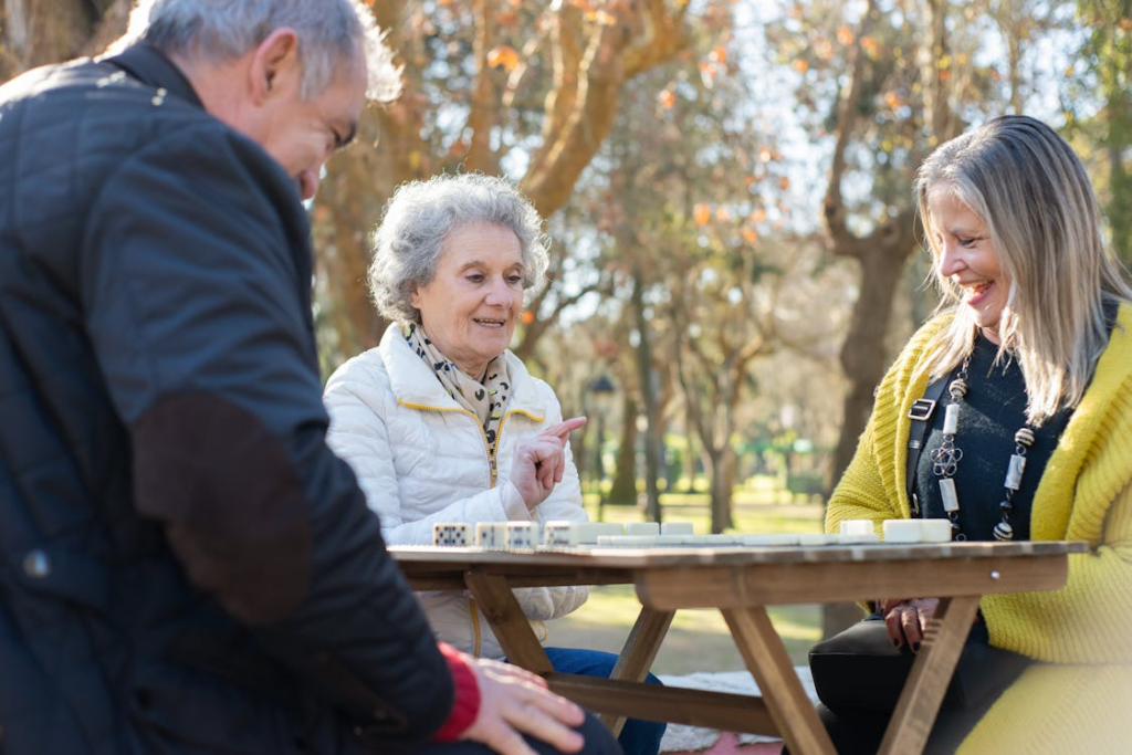 Three people playing dominoes outdoors on a wooden table. An elderly woman with gray hair wearing a white jacket is gesturing at the game while interacting with a middle-aged man and a woman with blonde hair in a bright yellow cardigan. Trees with autumn-colored foliage can be seen in the background on a sunny day.