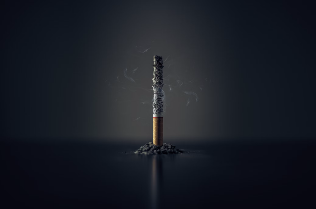 A close-up photograph of a lit cigarette standing vertically against a black background. The cigarette has a long ash column and thin streams of smoke rising from it. The image uses dramatic lighting to highlight the cigarette against the dark setting. Avoiding smoking is one important way to lower dementia risk.