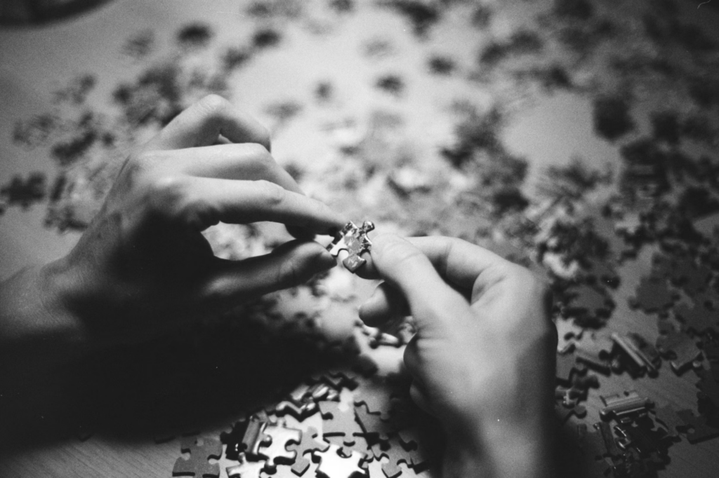 A black and white close-up photograph of two hands working on a jigsaw puzzle. The hands are shown fitting puzzle pieces together, with numerous other puzzle pieces scattered across the surface in the background. The monochromatic style creates a contemplative mood. Puzzles represent activities that may help lower dementia risk through mental stimulation.