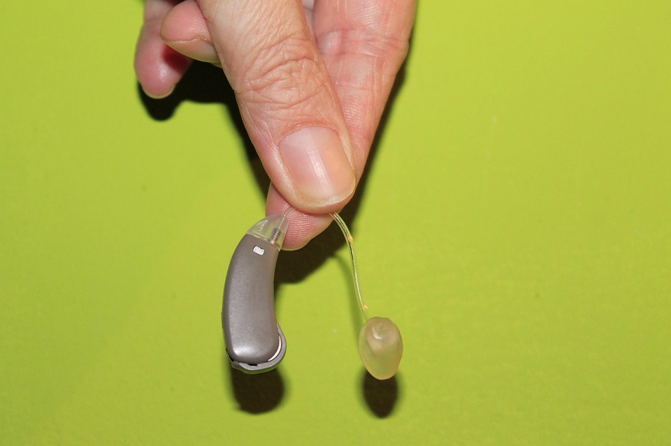 A close-up photograph of a person's thumb and finger holding a behind-the-ear hearing aid against a lime green background. The hearing aid is silver-colored with a clear plastic tube attachment. The image shows the details of the modern assistive listening device.