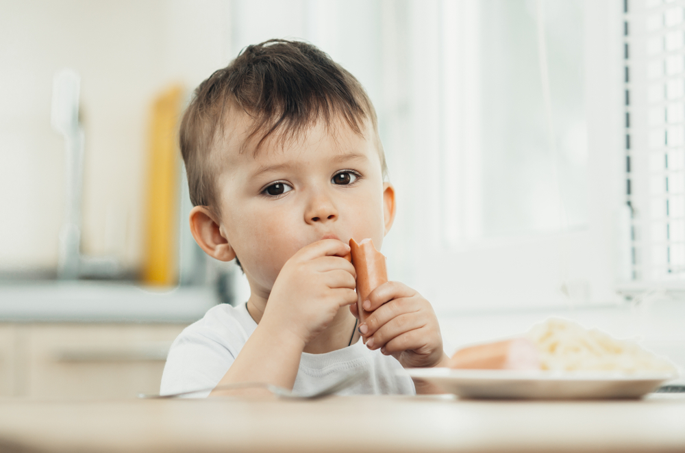 The child is sitting in the kitchen and greedily eats sausage
