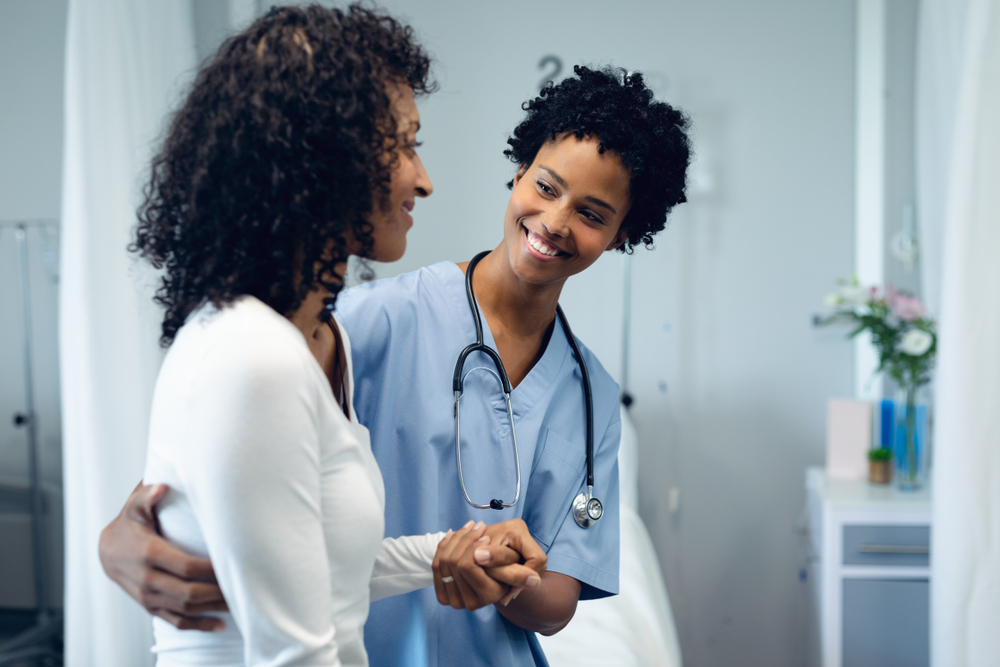 Front view of happy African American female doctor helping female patient to walk in the ward at hospital
