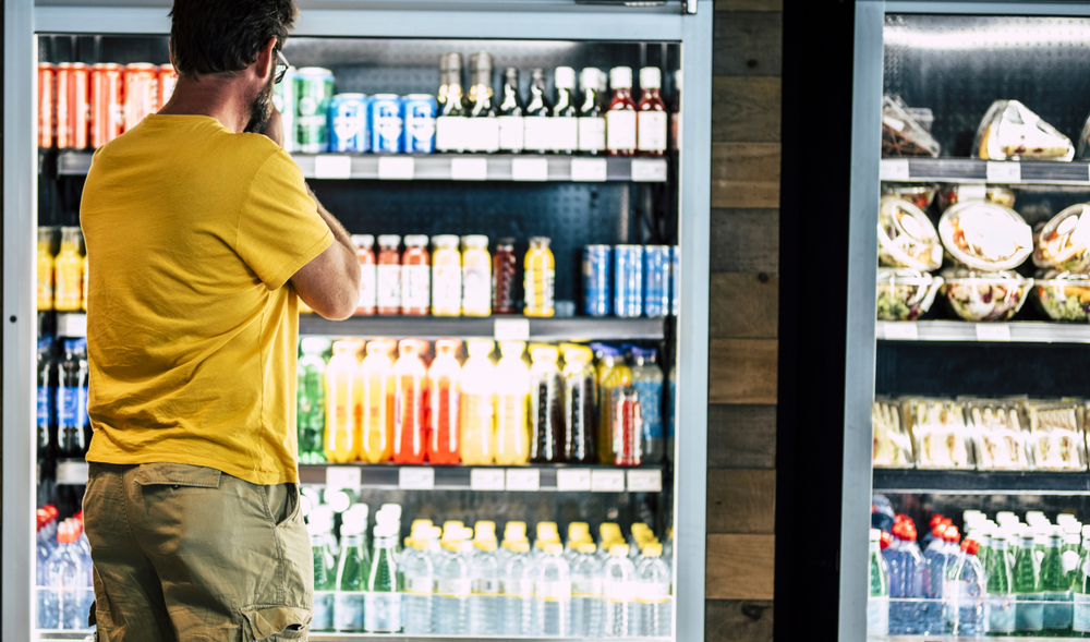man alone and isolated at the supermarket or minimarket choosing his drink or snack in big fridge with lots of products - difficult select and choose
