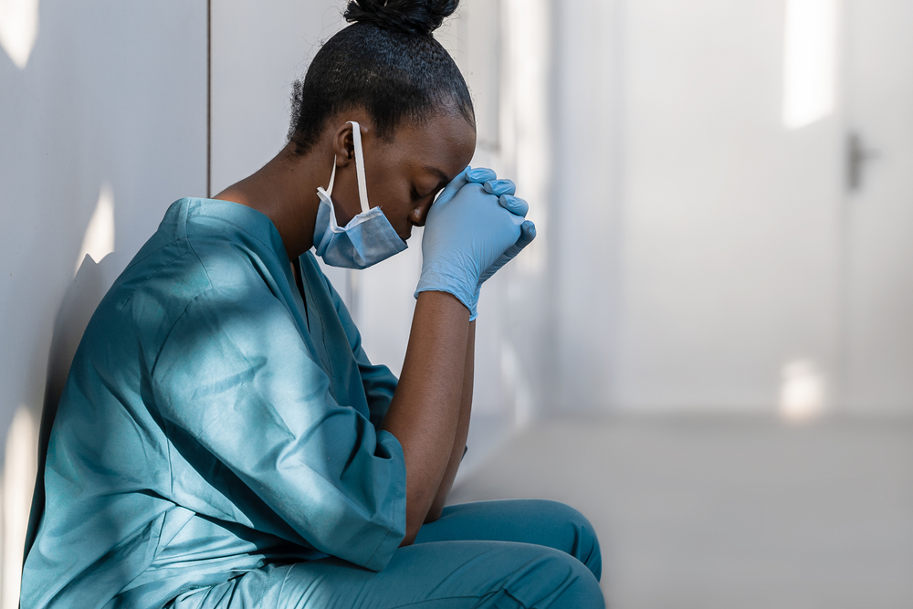 Tired depressed female african scrub nurse wears face mask blue uniform gloves sits on hospital floor. Exhausted sad black doctor feels burnout stress of corona virus frontline protection pray at work
