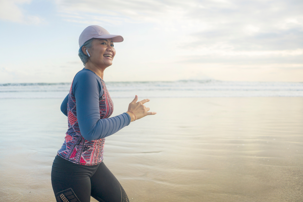 fit and happy middle aged woman running on the beach - 40s or 50s attractive mature lady with grey hair doing jogging workout enjoying fitness and healthy lifestyle at beautiful sea landscape
