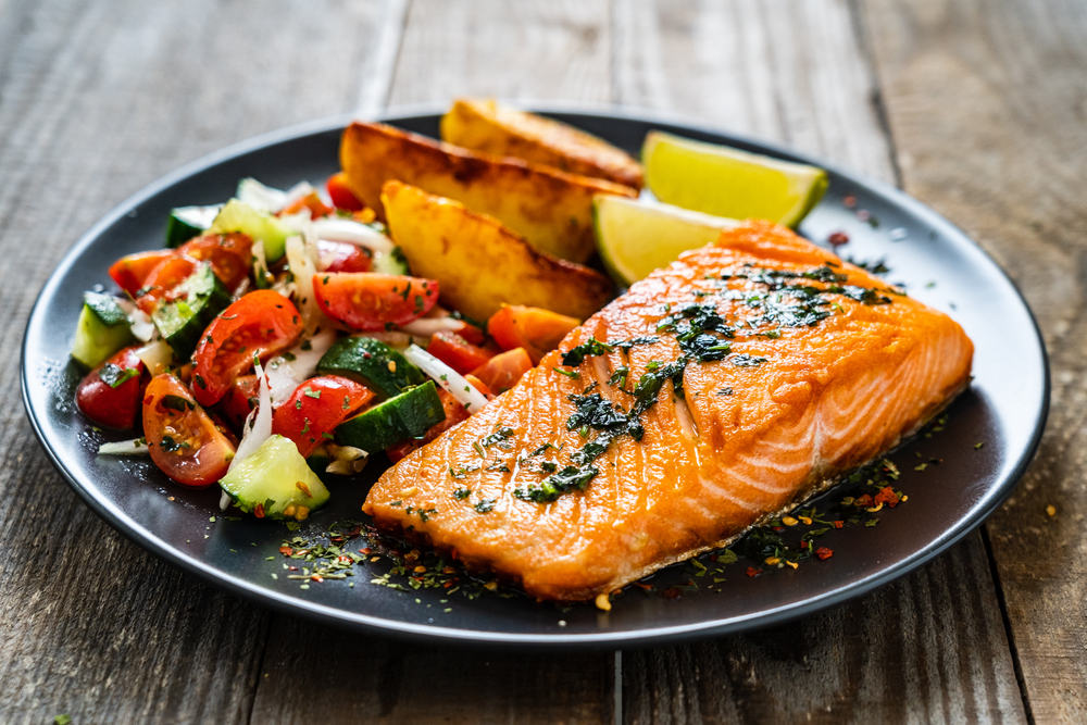 Fried salmon fillet with fried potatoes, lime and vegetable salad served on black plate on wooden table