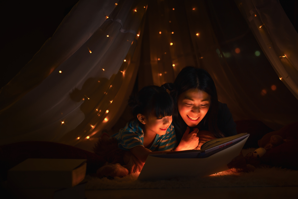 Asian family mom and girl happy at night time in bedroom at home before bed time reading fantasy bedtime story book together in child tent with dim yellow light
