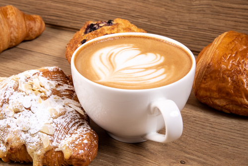 a white cup of cappuccino coffee with a heart shaped decorated foam with a selection of Danish pastry