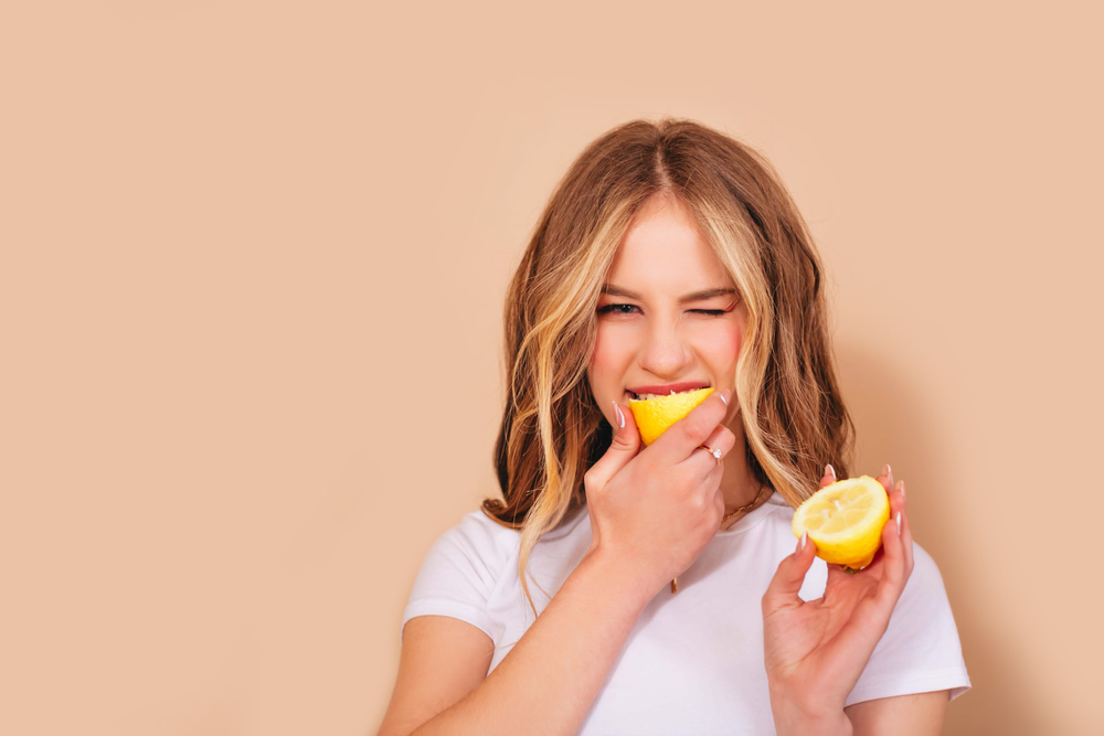 A teenage girl with wavy hair in a white t-shirt bites the lemon and grimaces in displeasure from the sour taste isolated on a beige background. Copy space.