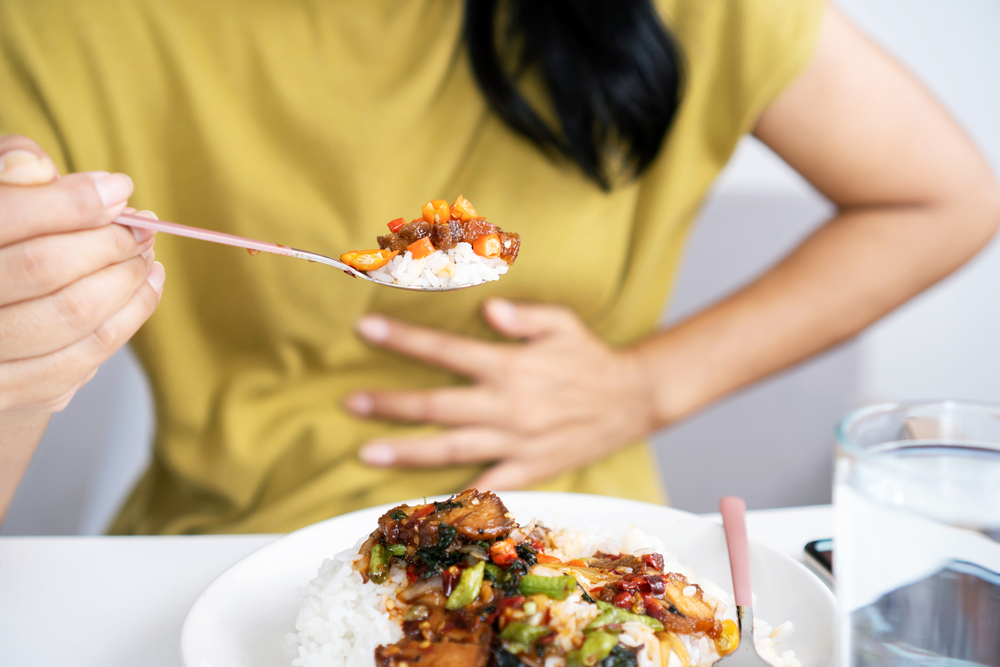 Asian woman eating spicy food and having acid reflux or heartburn hand holding a spoon with chili peppers another hand holding her stomach ache
