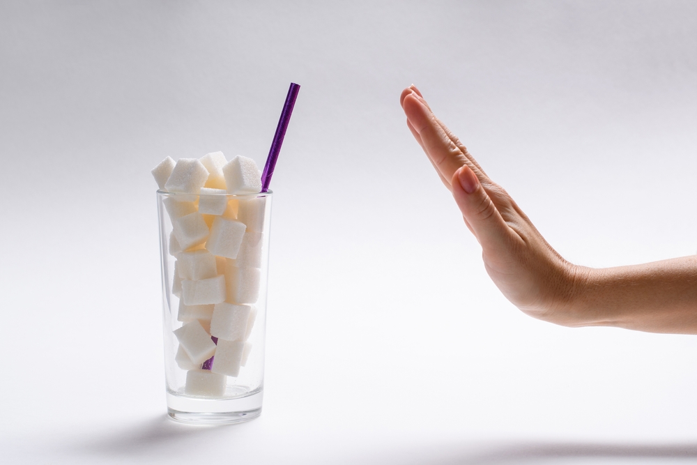 Stop sugar! A full glass of sugar and a woman's hand with a refusal gesture. The concept of avoiding the use of sugary drinks and cocktails. Selective focus on the glass.