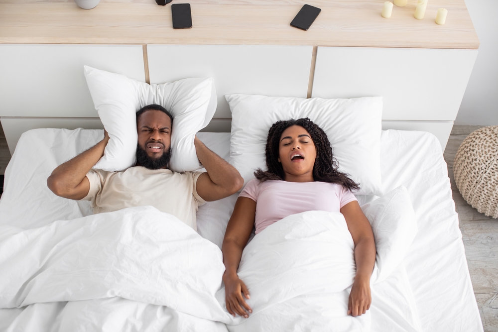 Angry disgruntled millennial black husband covers his ears with pillow, suffers from snoring and noise of wife at night on bed in bedroom interior, top view. Problems at home, insomnia and apnea