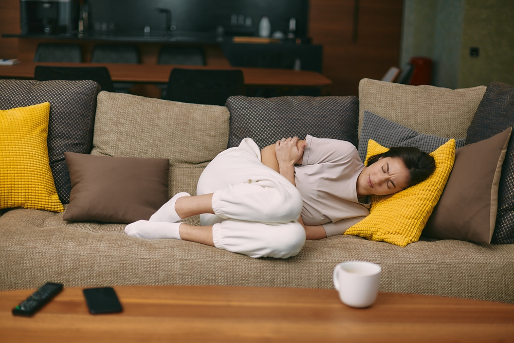 Sick young woman lying on sofa holding on stomach suffering abdominal pain from menstruation, diarrhea, or indigestion
