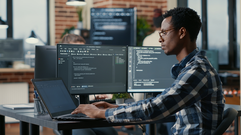 Portrait of african american developer using laptop to write code sitting at desk with multiple screens parsing algorithm in software agency. Coder working on user interface using portable computer.

