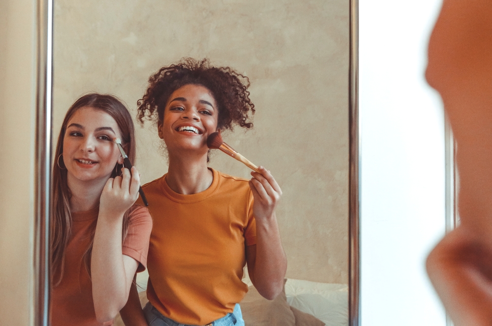 Female friendship. Two joyful multiracial girlfriends apply makeup with brush, having fun while standing in front of large mirror at home, female best friends of different races enjoy beauty routine
