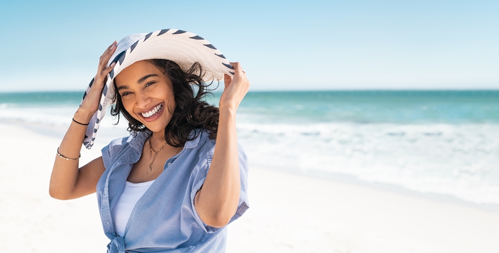 Portrait of stylish latin hispanic woman with white straw hat standing at beach. Young smiling woman on vacation enjoy sea breeze wearing straw hat and looking at camera. Attractive beautiful girl.
