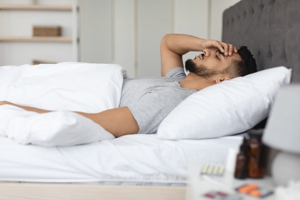 Side View Shot Of Sick Arab Man Lying In Bed At Home, Ill Middle Eastern Man Feeling Unwell And Touching Forehead While Resting In Bedroom, Having Medicine Pills On Bedside Table, Selective Focus