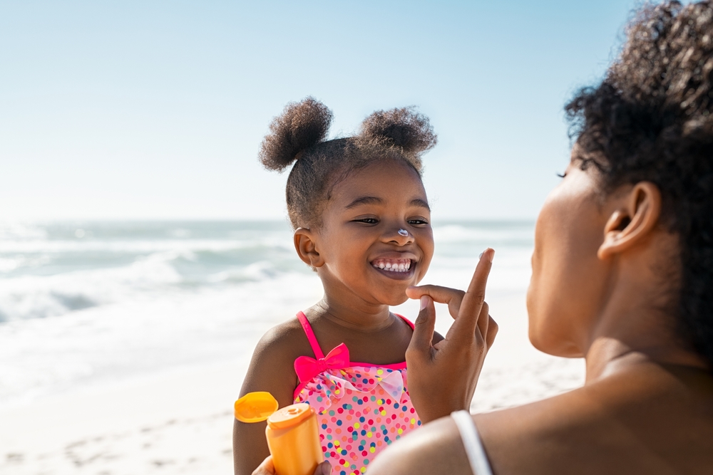 Young mother applying protective sunscreen on daughter nose at beach with copy space. Black woman hand putting sun lotion on female child face. African american cute little girl with sunblock cream.
