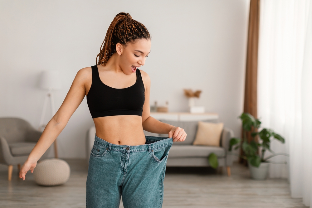 Excited Slim Black Female After Great Weight Loss Showing Result Of Slimming Wearing Old Big Jeans Standing Indoors. Successful Dieting And Slimming Concept