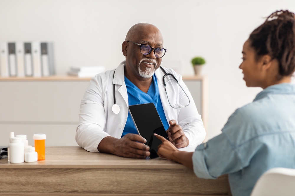 Medicine And Health Care Concept. Black male doctor in glasses holding digital tablet with empty screen for mockup, showing female patient her test results and discussing treatment, sitting at desk
