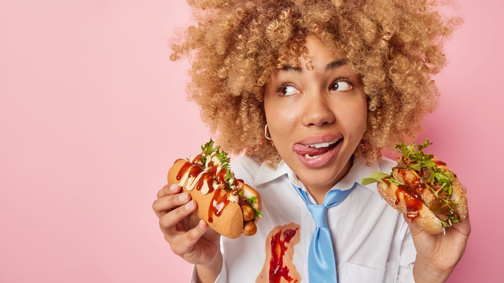 Horizontal shot of thoughtful curly haired woman licks lips chooses between hot dog and burger eats unhealthy food dressed in formal clothes isolated over pink background blank space for your text
