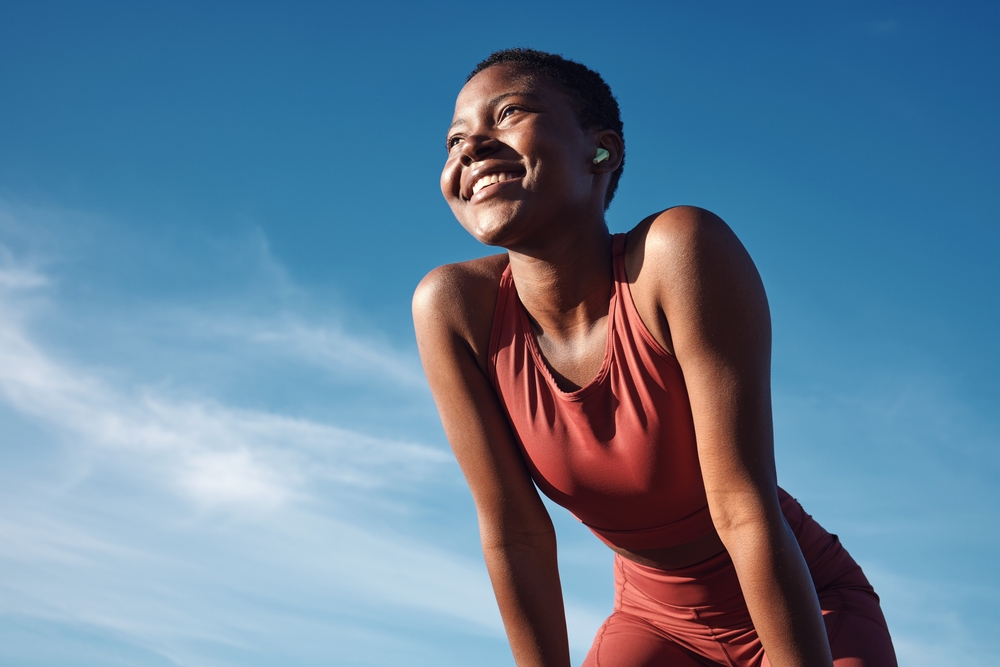 Fitness, black woman and happy athlete smile after running, exercise and marathon training workout. Blue sky, summer sports and run of a African runner breathing with happiness from sport outdoor
