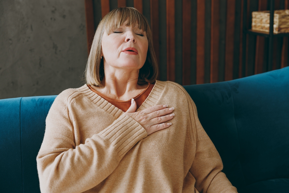 Close up elderly ill sad woman 50s year old wear casual clothes sits on blue sofa put folded hands on heart suffer from pain close eyes stay at home flat spend free spare time in living room indoor
