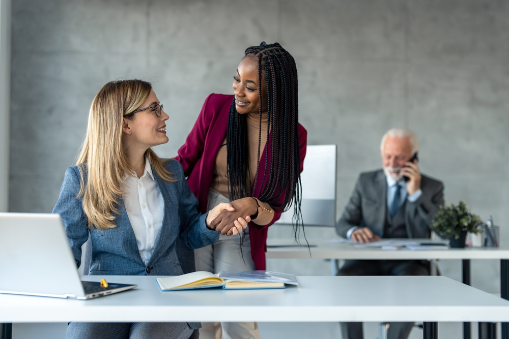 Two cheerful multicultural professional business women female employees in suit supporting each other at work place, holding hands and smiling. Business team cooperation concept.
