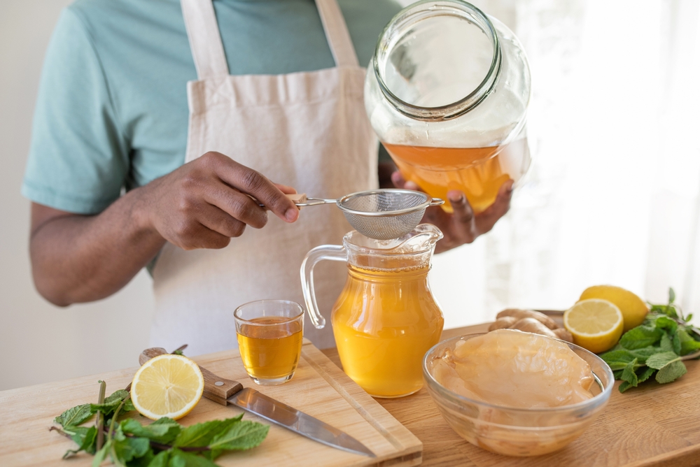 Making kombucha tea drink at home, fermented drink flavoured with ginger, lemon and mint. Healthy drink, organic probiotic beverage. Black young male hands pouring drink through a sieve.
