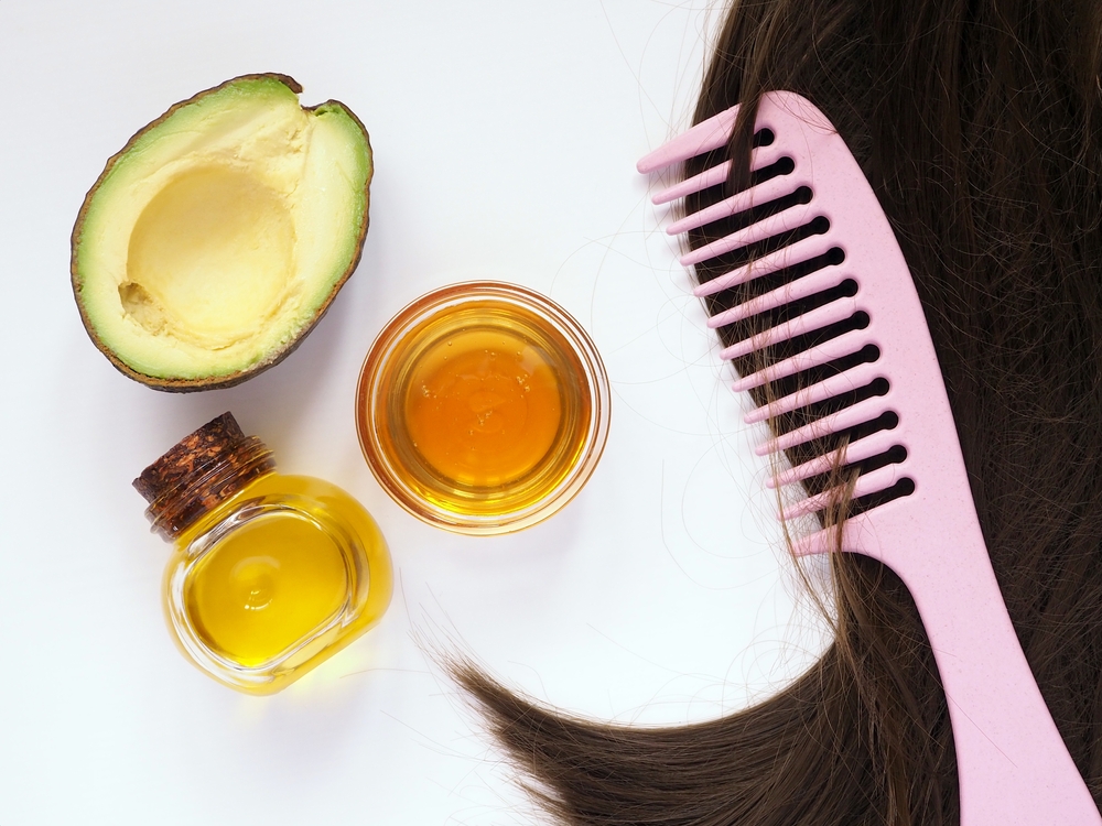 Ingredients for homemade hair mask: cosmetic oil, honey, avocado, brown hair strand and comb. Isolated on white background. Flat lay.