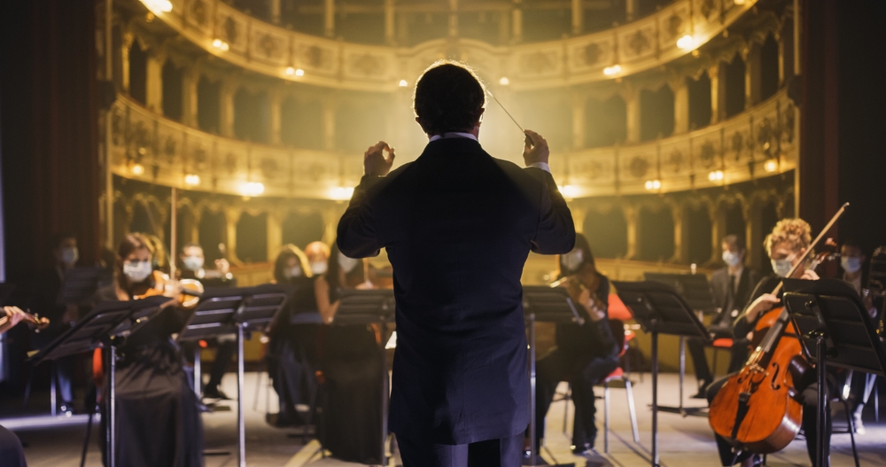 Back View of Professional Conductor Directing Symphony Orchestra with Performers Wearing Medical Masks, Playing Violins, Cello and Trumpet on Classic Theatre with Curtain Stage During Music Concert

