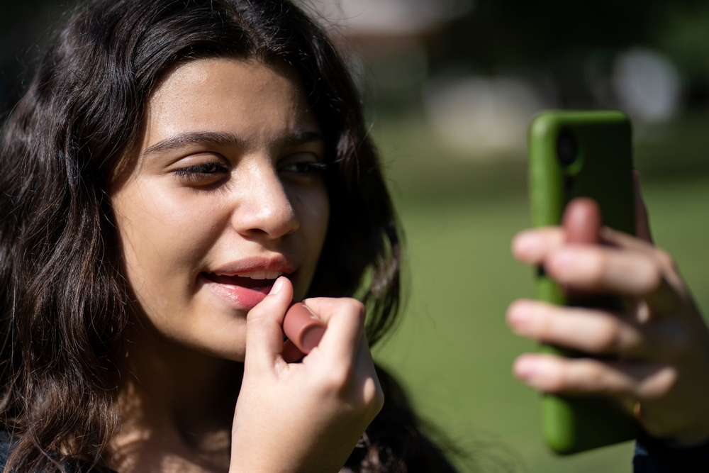 Latina teen in a park putting on lipstick using her phone as a mirror
