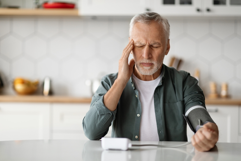 Hypertension Concept. Senior Man Checking Blood Pressure With Upper Arm Monitor At Home, Elderly Gentleman Touching Temples And Measuring Arterial Tension While Sittin At Table In Kitchen, Copy Space
