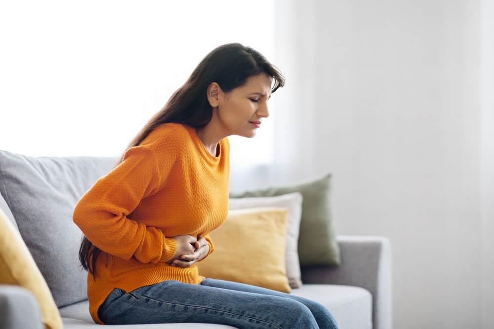 Unhappy sick pretty long-haired young indian woman wearing casual comfy outfit sitting on couch in living-room at home, touching belly, suffering from period crumps, side view, copy space