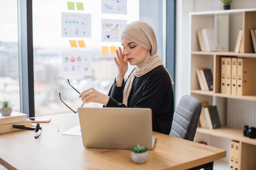 Overworked arabian woman in hijab massaging nose after taking off glasses while sitting at writing desk in office. Weary business person feeling constantly exhausted because of overtime job.

